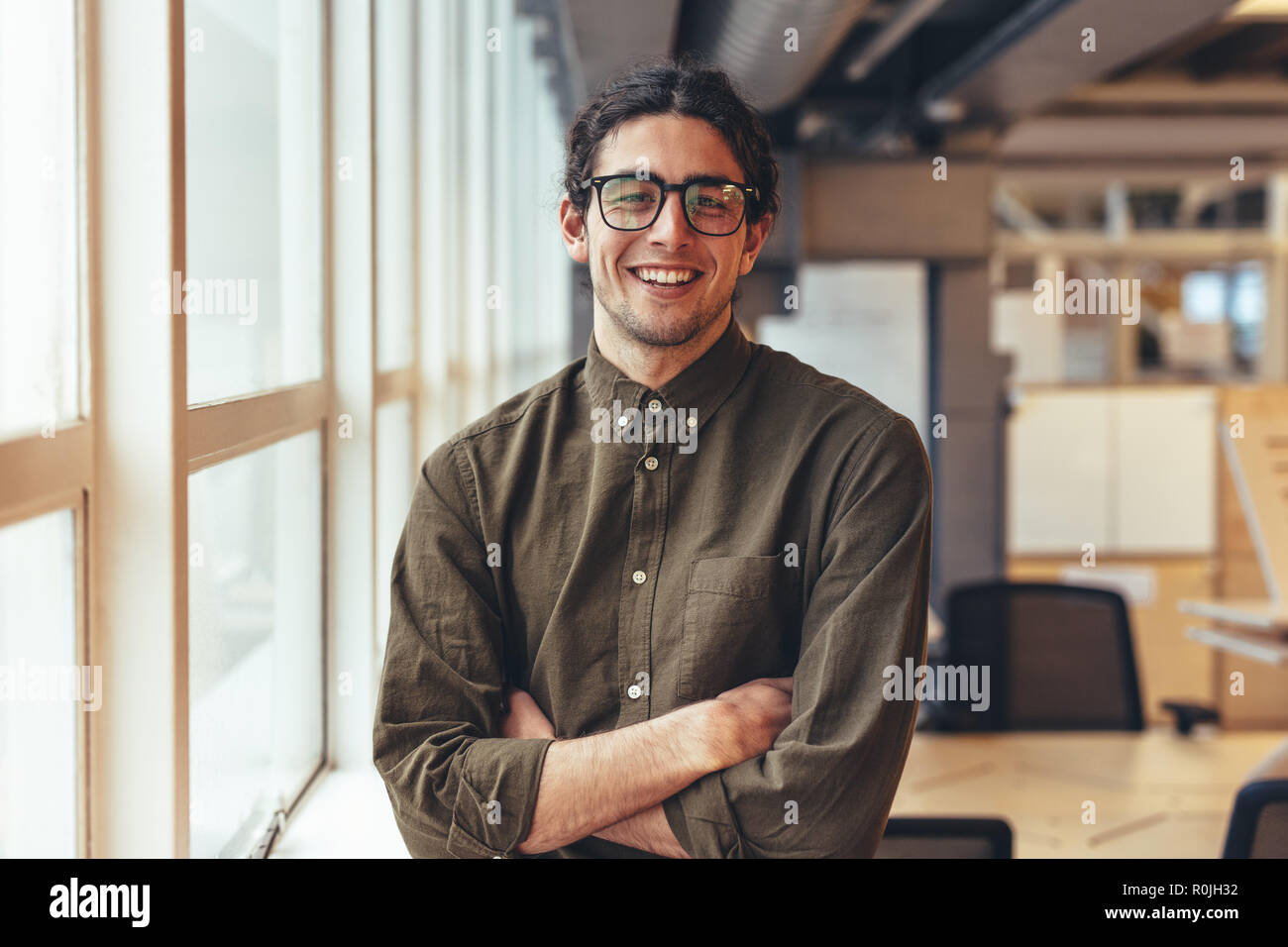 Businessman standing beside a window in office with arms crossed. Portrait of a smiling man standing in office beside a window. Stock Photo