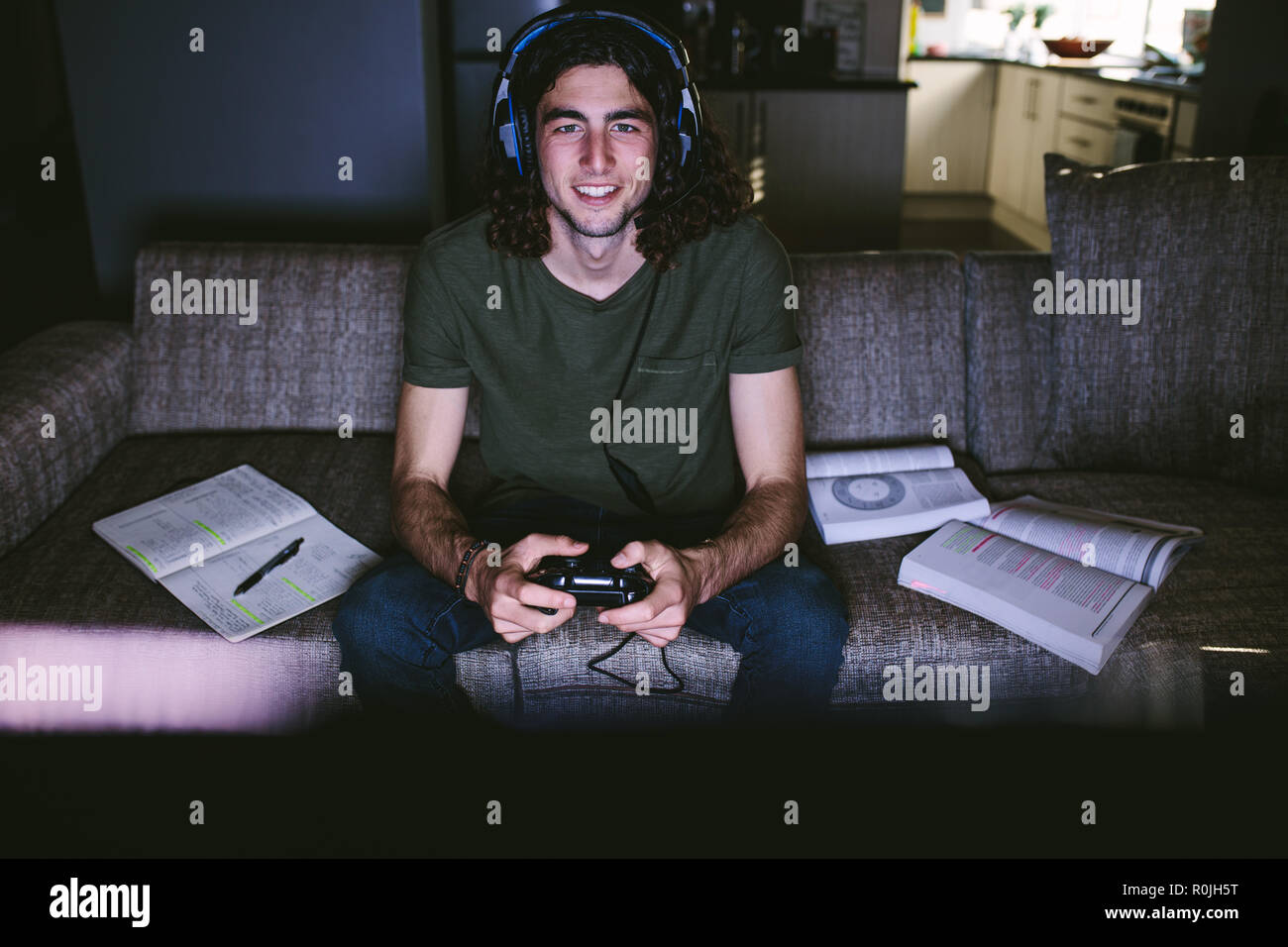 Student sitting on couch playing video game with books beside him. Student taking a break from studies playing a game. Stock Photo