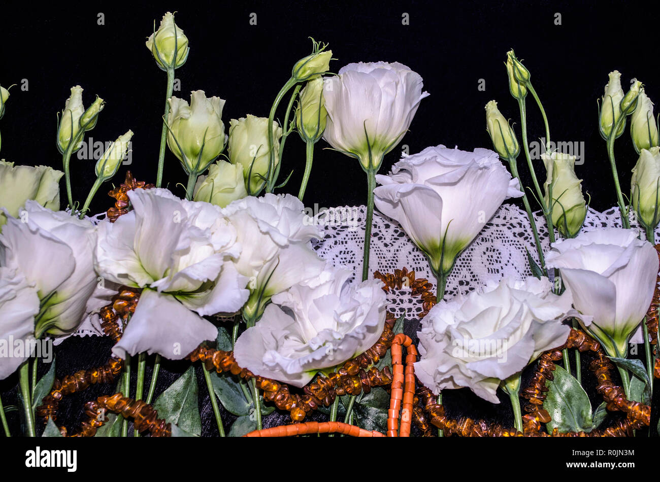 White flowers of Lisianthus with red coral and amber beads on a white openwork embroidered edging,lie the black plywood Stock Photo