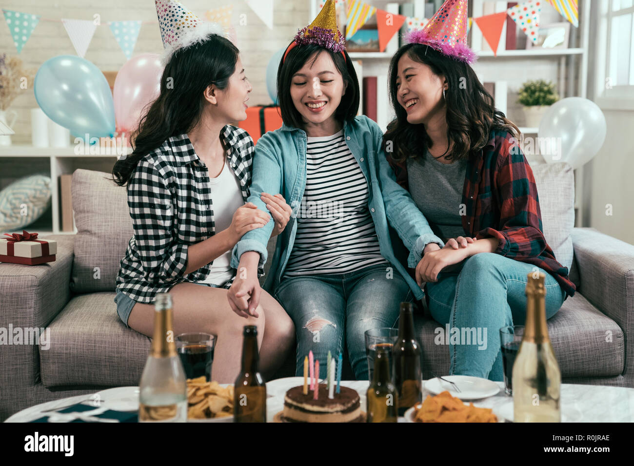 Group of young friends with food and bottles of drink celebrating in home interior. sisters having fun birthday house party with funny hats. girls sit Stock Photo