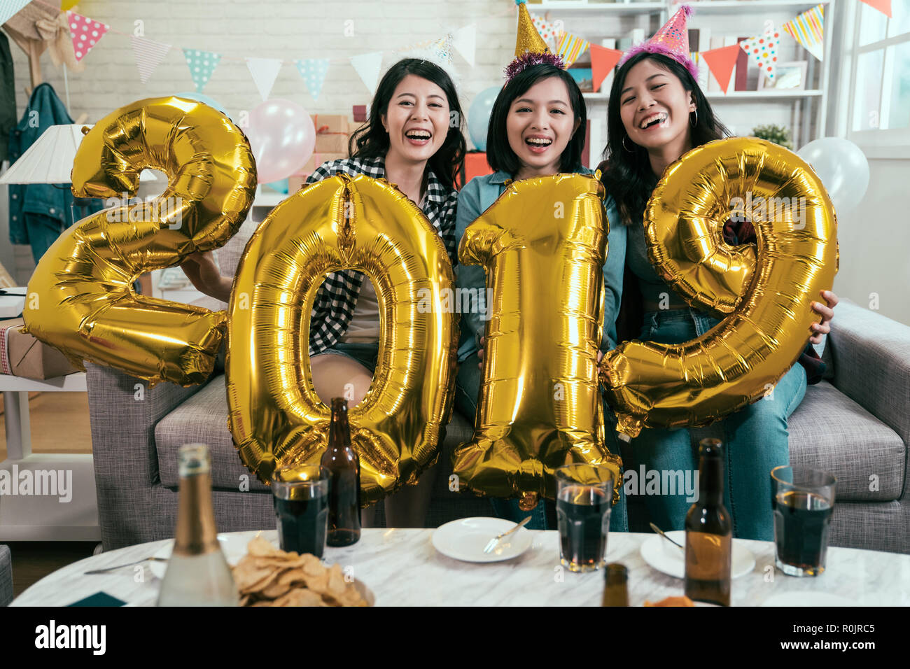 friends carrying gold 2019 number in house party celebrating new year. young girls enjoy chilling at home with drinks beer and food on table. asian fe Stock Photo