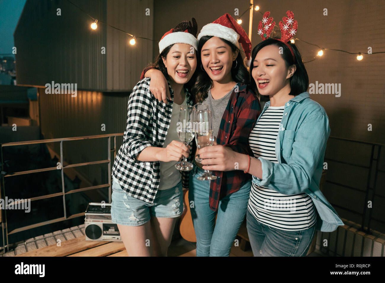 dark night christmas party outdoor. cheerful ladies with santa hats cheers champagne on balcony in xmas eve. young asian girls celebrate with music. Stock Photo
