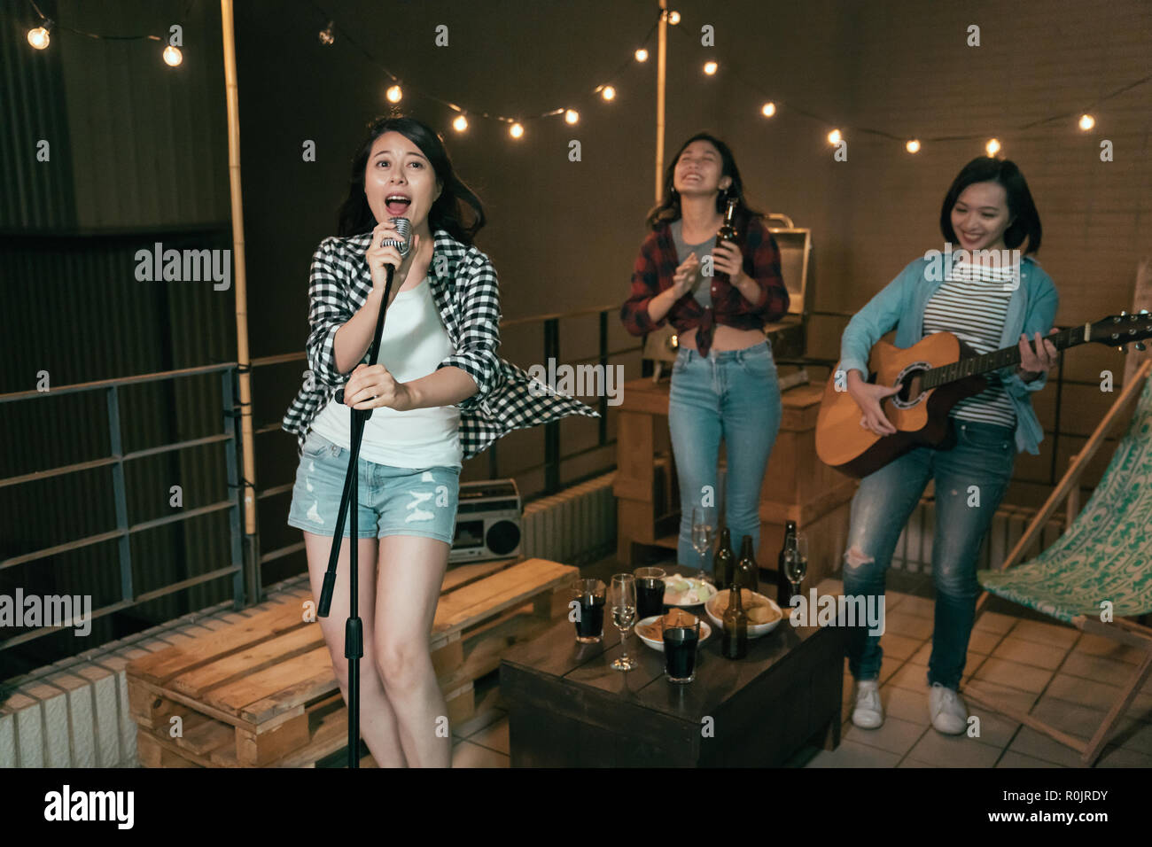 asian girl bands practicing new songs on the roof at night. young ladies singing playing guitar and dancing dark outdoor. crazy women on the balcony r Stock Photo