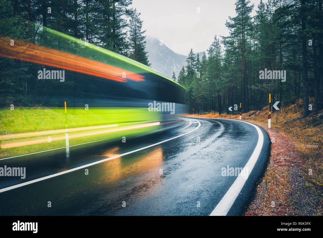 Blurred green bus on the road in autumn forest in rain. Perfect asphalt mountain road in overcast rainy day. Roadway, pine trees in alps. Transportati Stock Photo