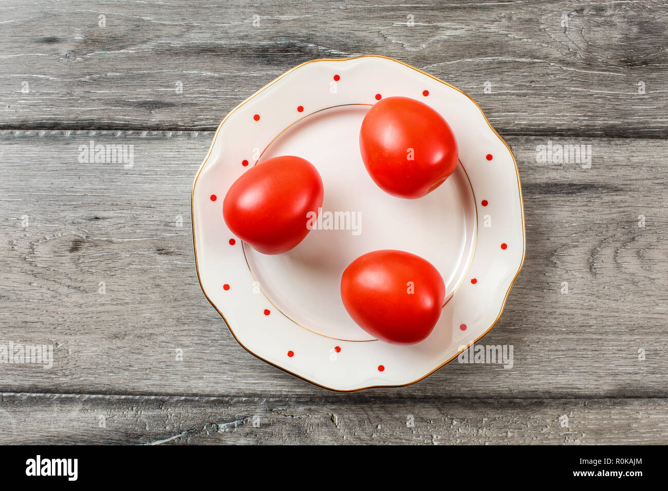 Three whole tomatoes on white plate with small red dots and golden rim, gray wooden desk, tabletop view. Stock Photo