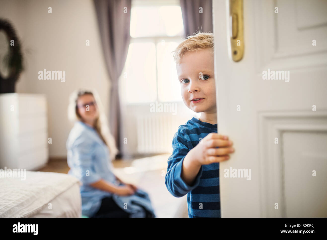 A toddler boy with young mother in the background inside in a bedroom. Stock Photo