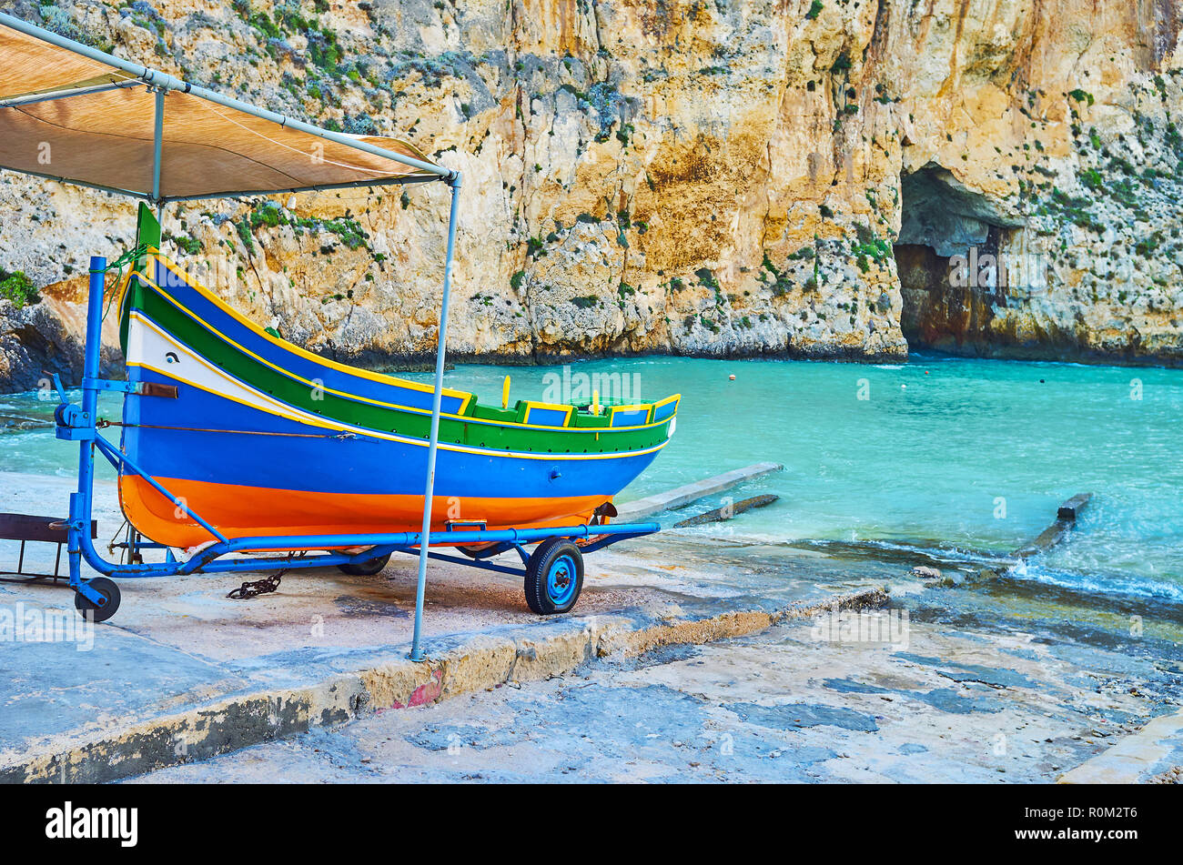 The colored wooden Maltese luzzu boat on shore of Dwejra Inland sea with a view on Blue Hole grotto on the background, San Lawrenz, Gozo, Malta. Stock Photo