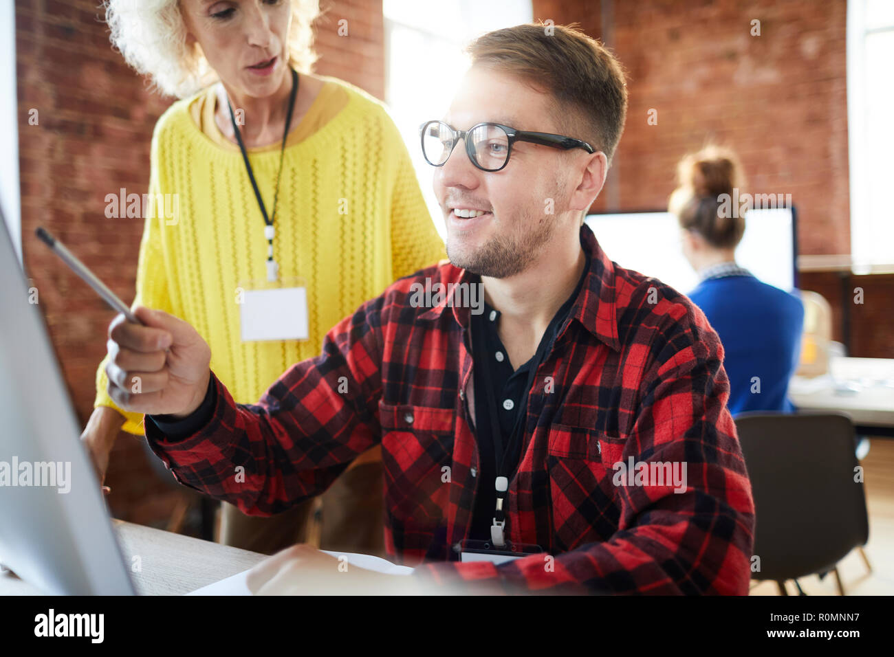 Young smiling analyst pointing at data on computer screen while explaining it to mature employer Stock Photo