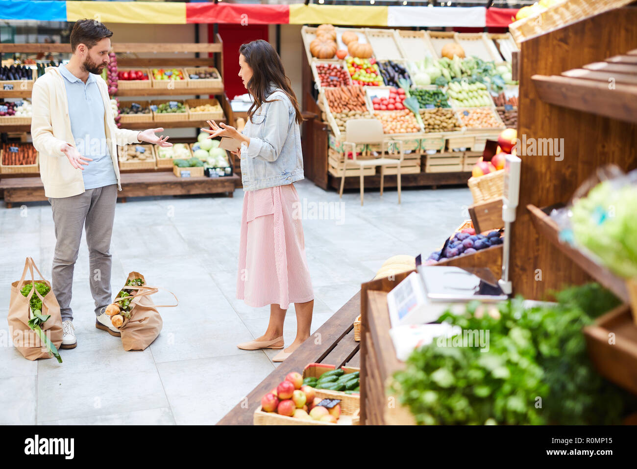 Outraged young couple in casual clothing standing in healthy food shop and gesturing while   fighting, they confronting with each other, shopping bags Stock Photo