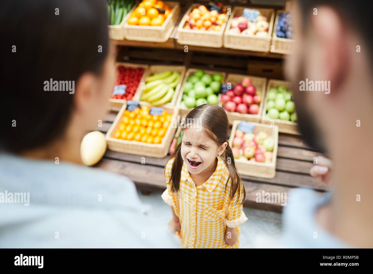 Upset hysterical girl with closed eyes crying loudly while manipulating parents and standing against food stall in supermarket Stock Photo