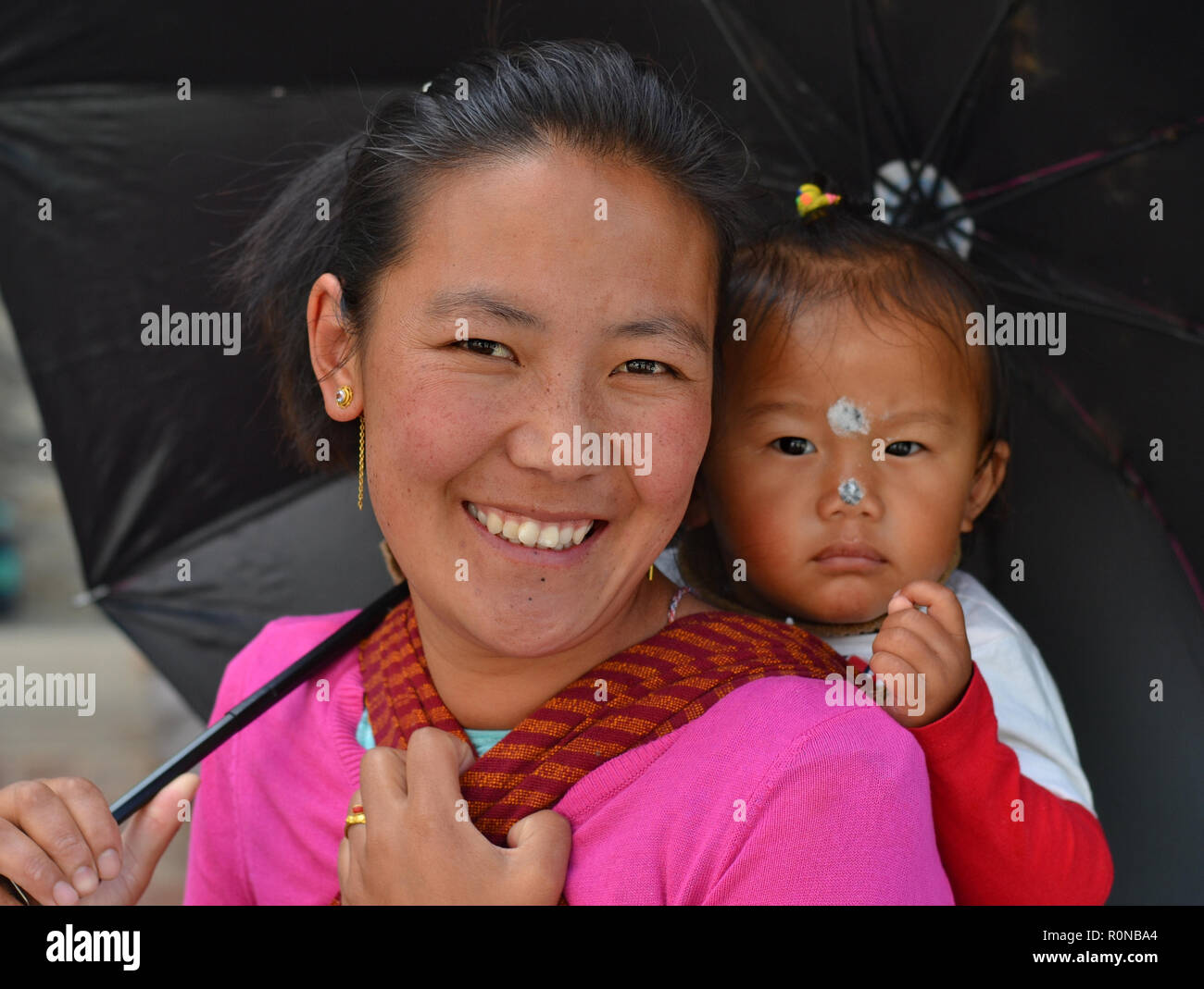 Young Indian Monpa hill-tribe woman carries her toddler girl in a traditional baby sling on her back. Stock Photo