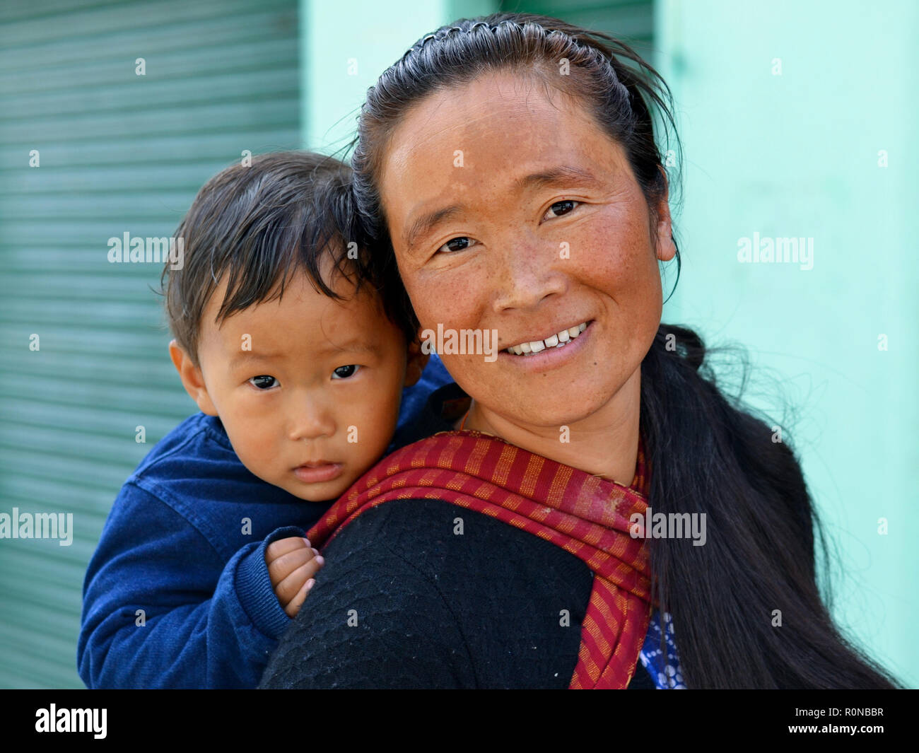 Young Indian Monpa woman carries her toddler boy in a traditional baby sling on her back. Stock Photo