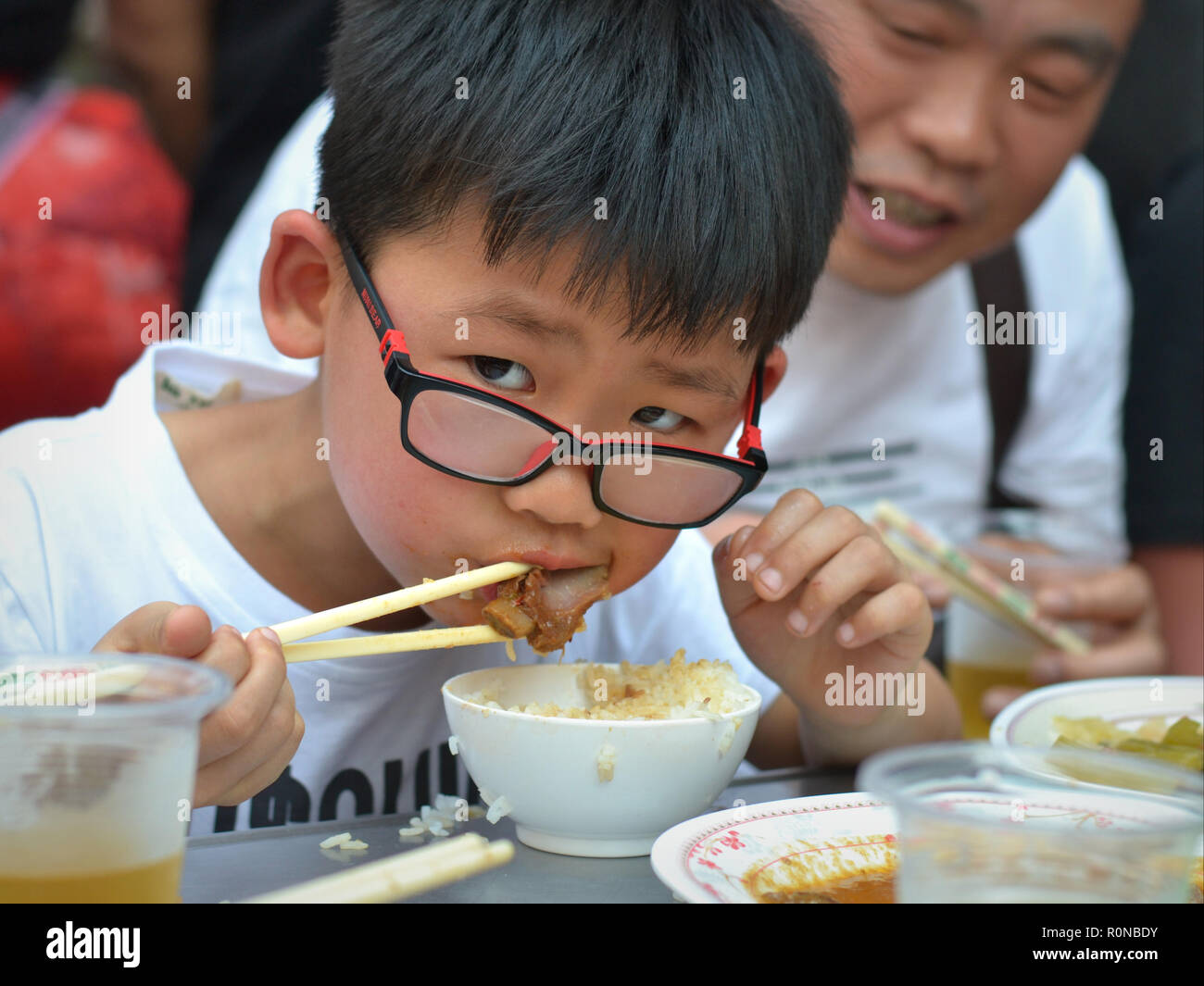 Little Chinese boy with nerdy eyeglasses eats steamed rice and pork meat with wooden chopsticks. Stock Photo