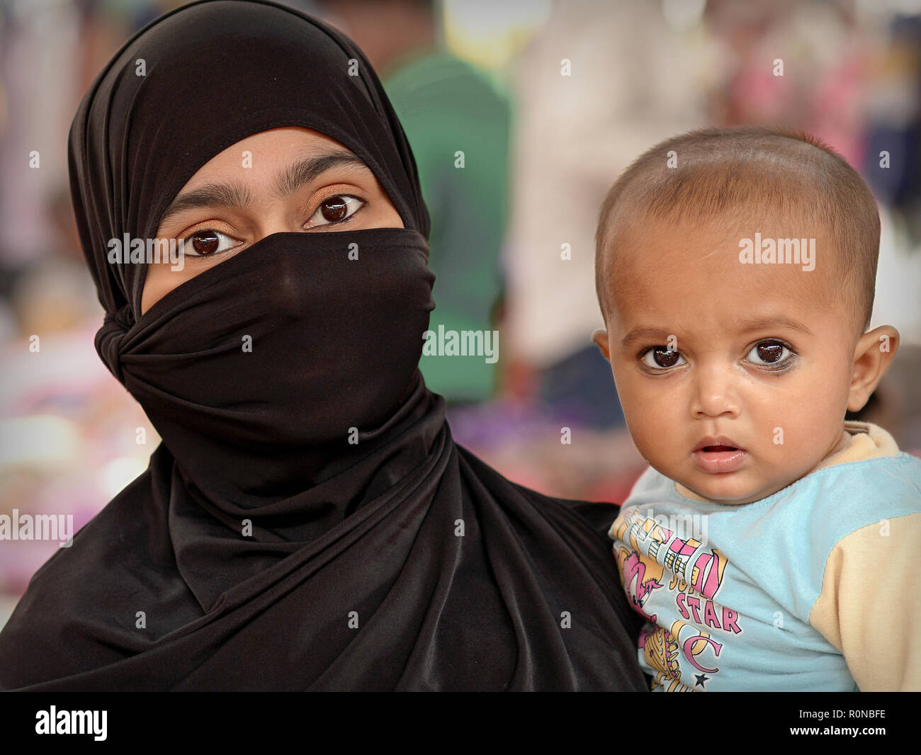 Young Indian Muslima with beautiful, almond-shaped eyes wears a black niqab and burqa and carries her little son in her arms. Stock Photo
