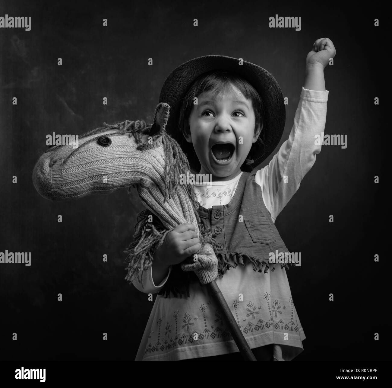 Screaming little girl with a homemade toy. Little girl dressed like a cowboy playing with a homemade horse. Expressive facial expressions. Black and w Stock Photo