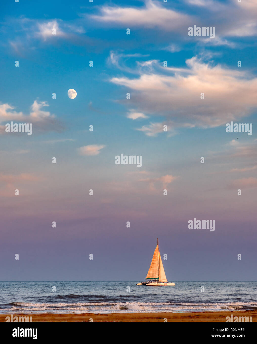 A sailboat on the sea with the nearly full moon in the sky above which is blue, pink and purple from the sun setting Stock Photo