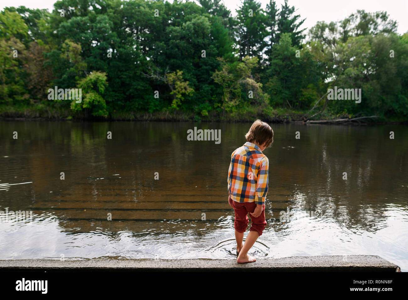 Boy standing by a river dipping his toe in the water, United States Stock Photo