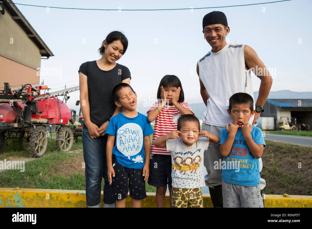 Portrait of smiling Japanese farmer, his wife and four children standing in their yard, children pulling faces at camera. Stock Photo