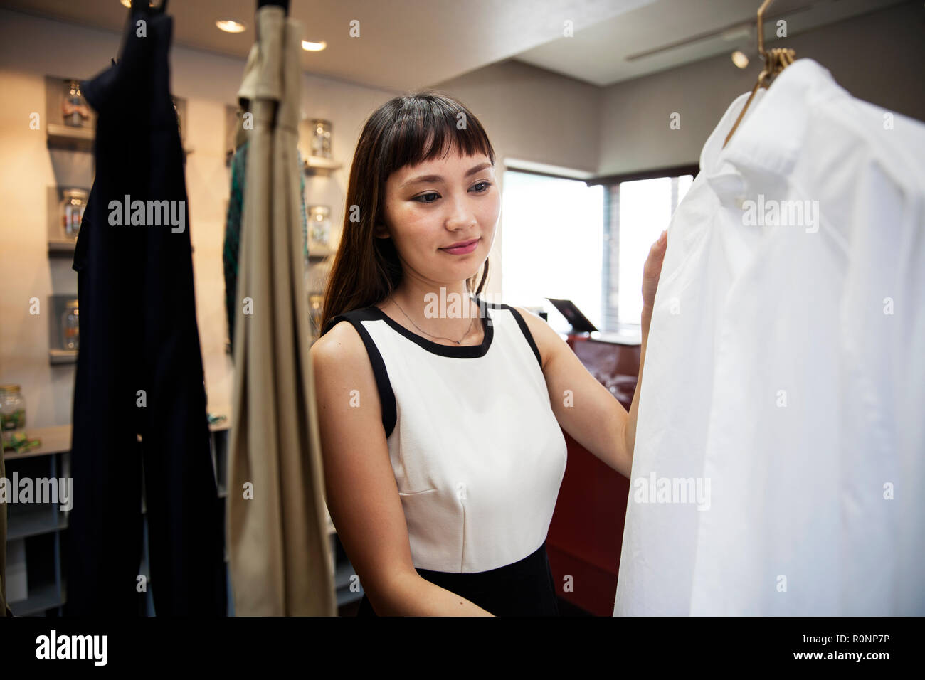 Smiling Japanese saleswoman standing in clothing store, looking at shirt. Stock Photo