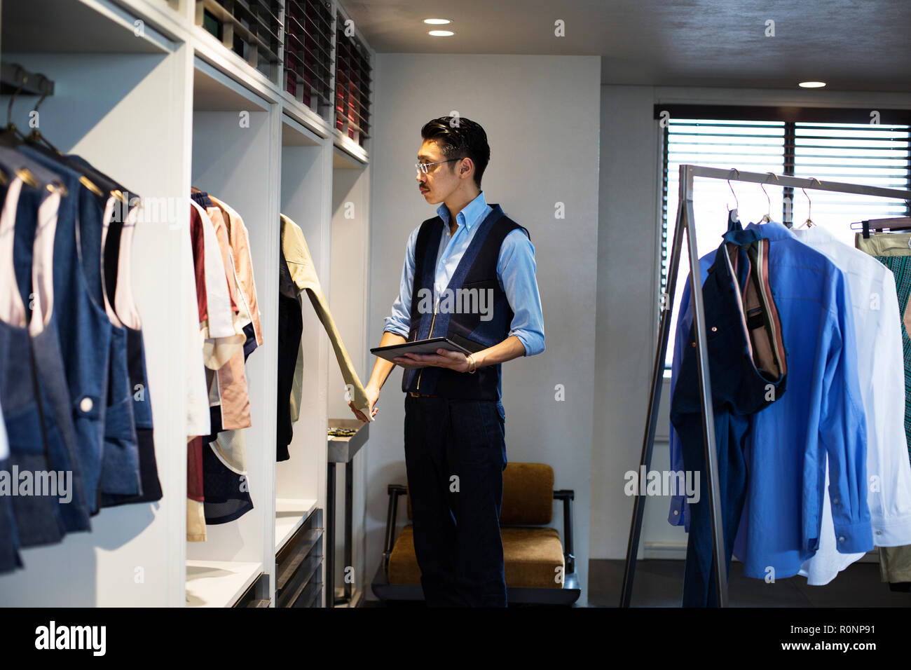 Japanese salesman with moustache wearing glasses standing in clothing store, looking at clothes on a rail, holding digital tablet. Stock Photo
