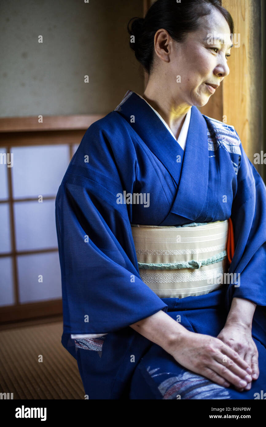 Japanese woman wearing traditional bright blue kimono with cream coloured obi kneeling on floor in traditional Japanese house. Stock Photo