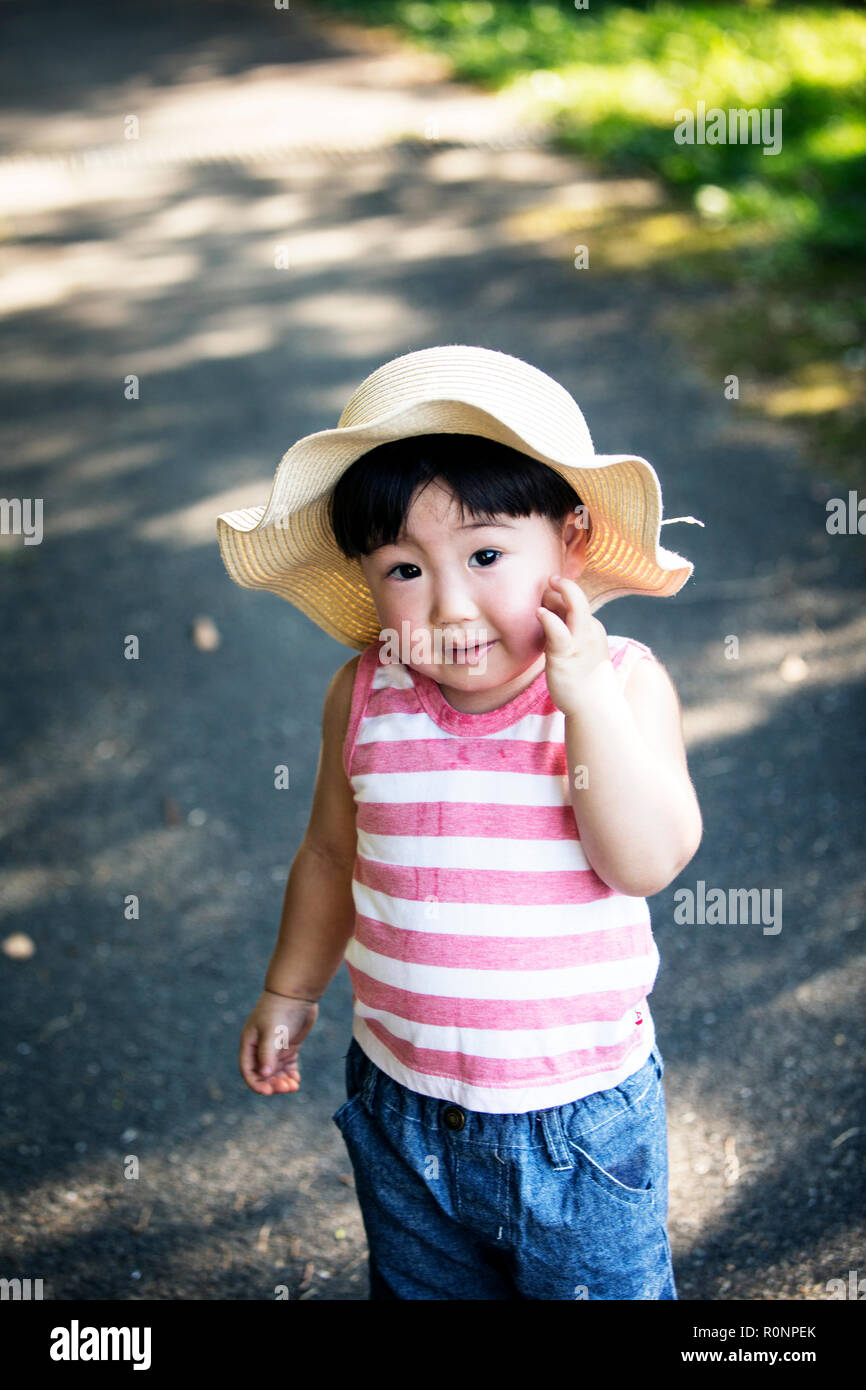 A cute toddler in a sunhat standing on a path outdoors in summer looking at camera hand on face Stock Photo