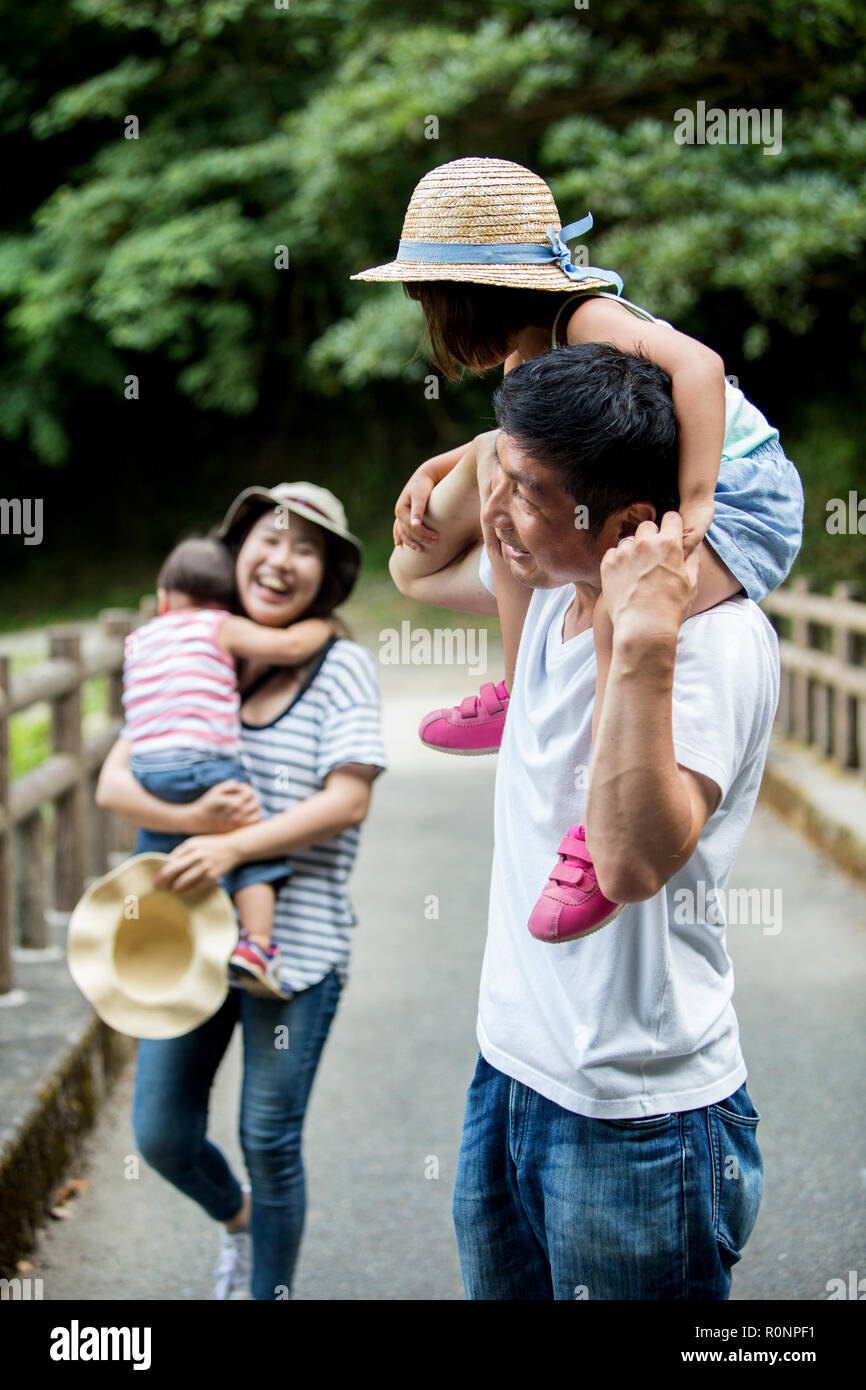 Japanese girl, smiling woman holding hat and man carrying toddler on his shoulders standing on wooden bridge. Stock Photo
