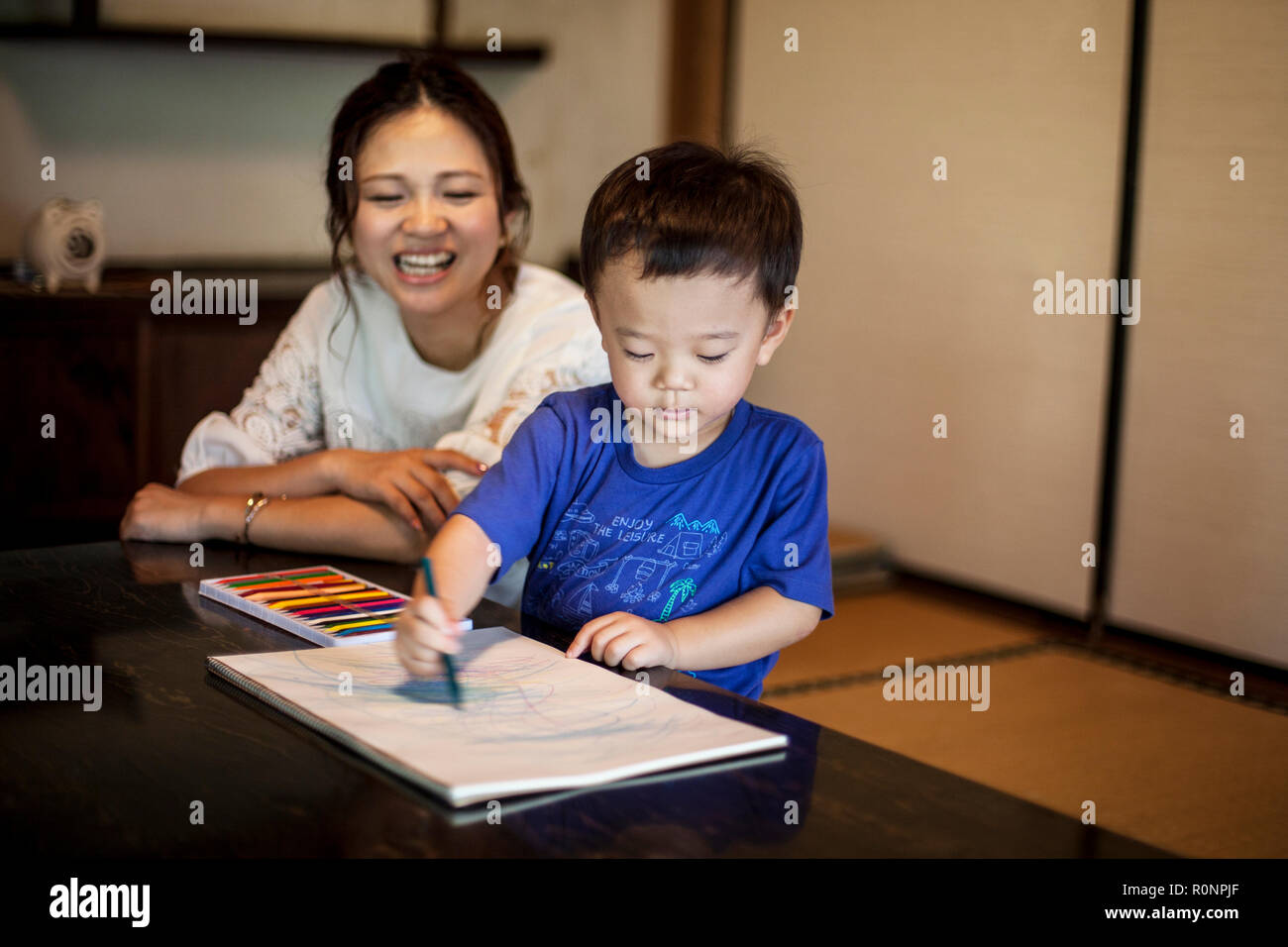 Smiling Japanese woman and little boy sitting at a table, drawing on white paper with colouring pens. Stock Photo