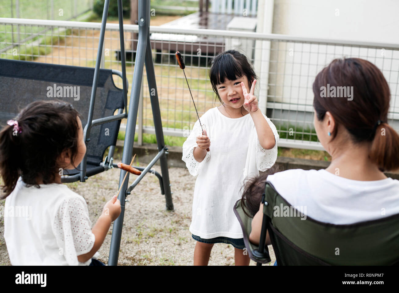 Two Japanese girls holding sausages on skewers and woman in a backyard. Stock Photo
