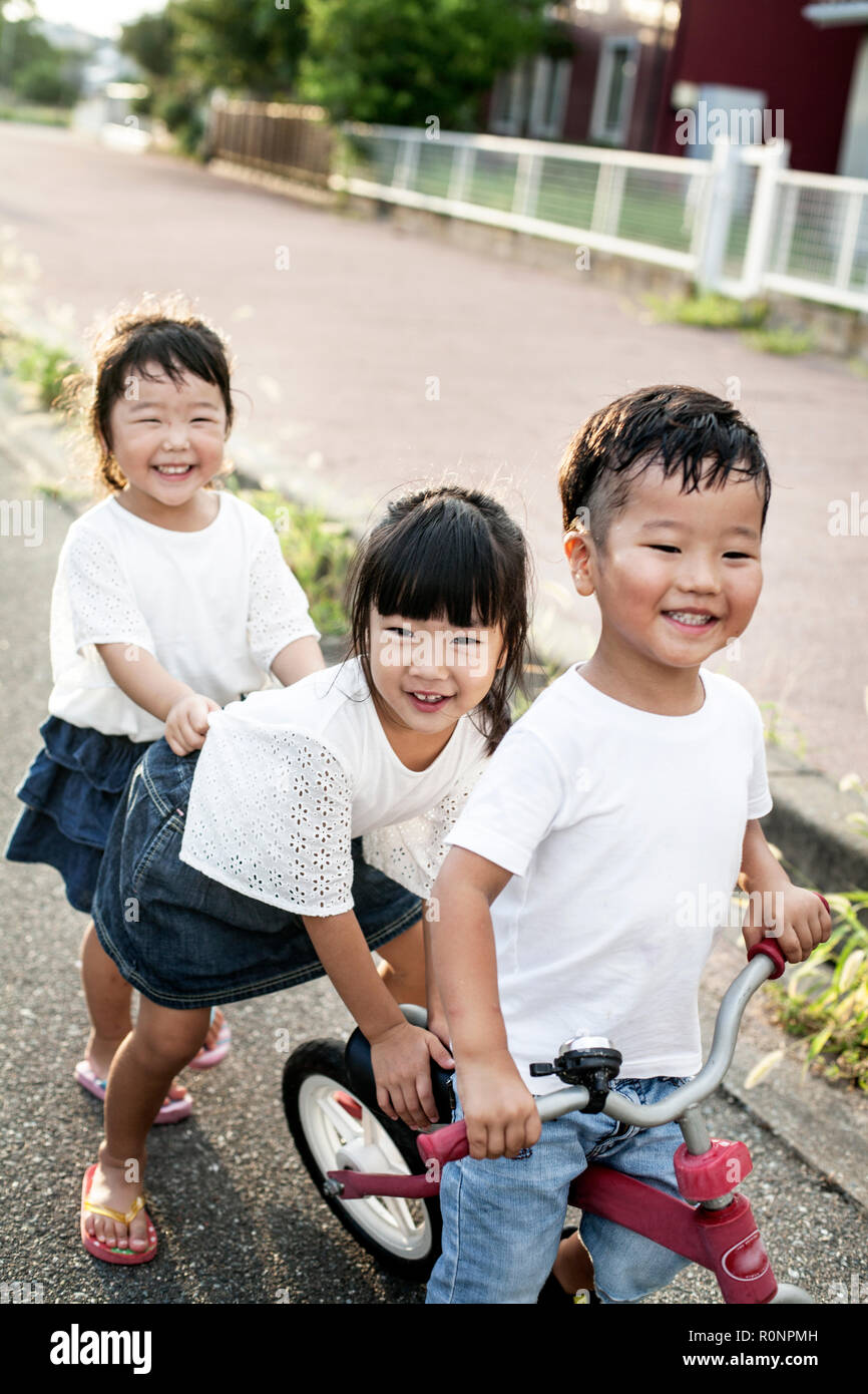 Portrait of two Japanese girls and boy playing on street with a bicycle, smiling at camera. Stock Photo