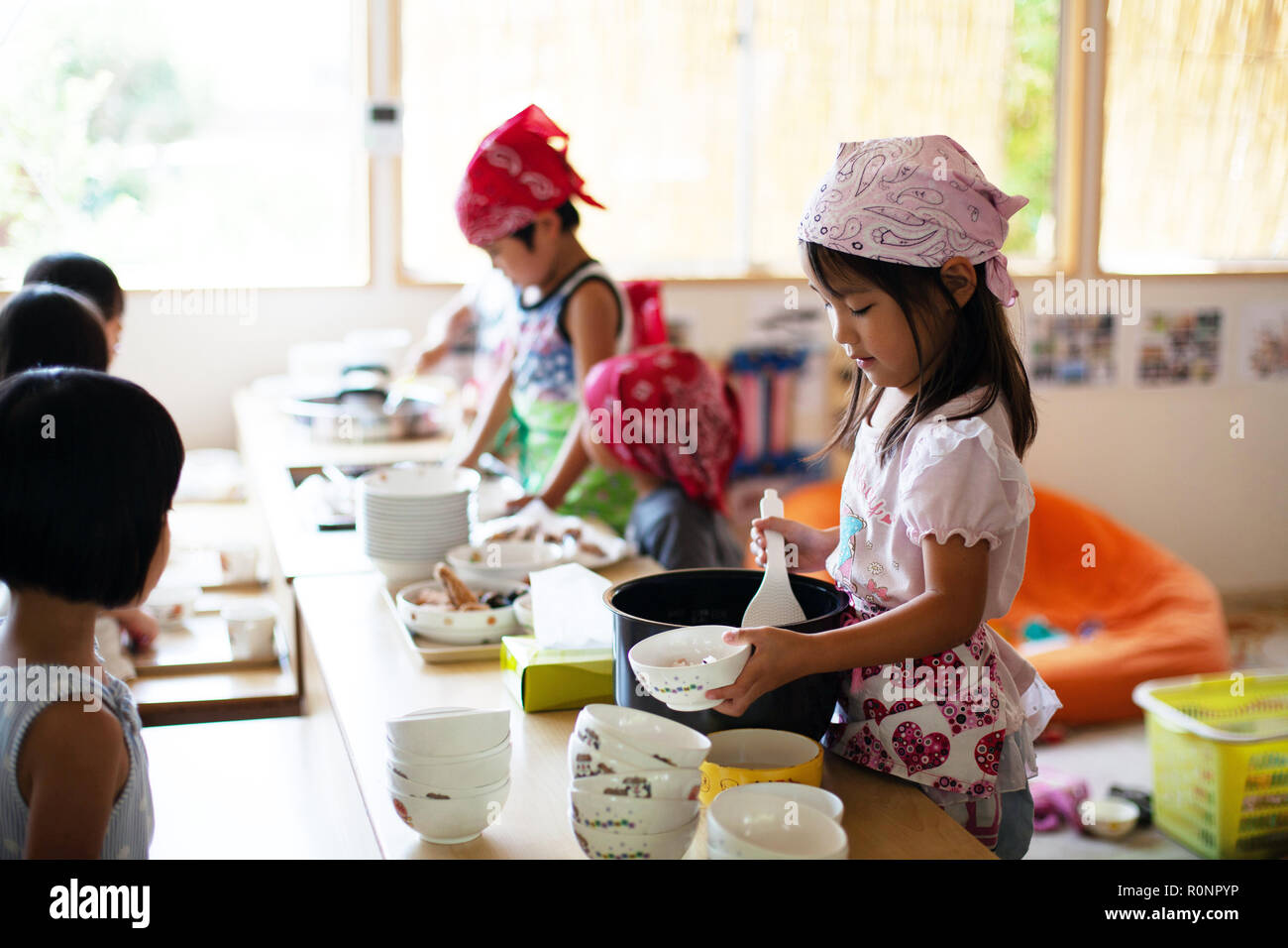A girl and a boy wearing headscarves standing at a table in a Japanese preschool, serving lunch. Stock Photo