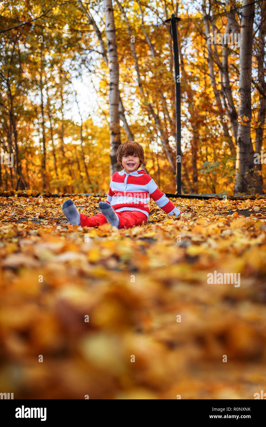 Boy sitting on a trampoline covered in autumn leaves, United States Stock Photo