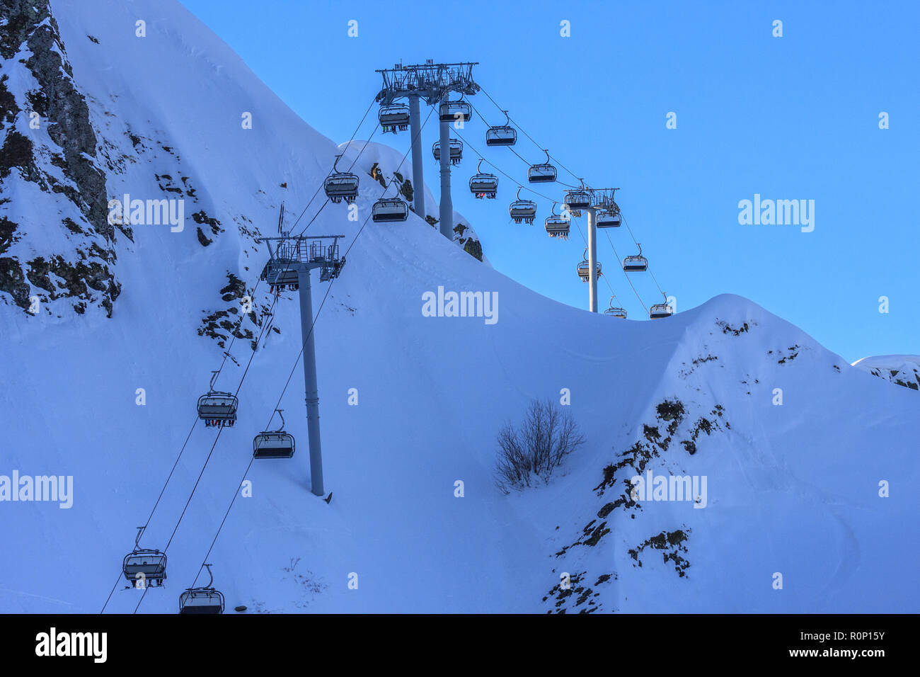 Skiers and snowboarders ride on chair ski lift at Gorky Gorod mountain ski resort in Sochi, Russia, against blue sky in the evening of sunny winter da Stock Photo