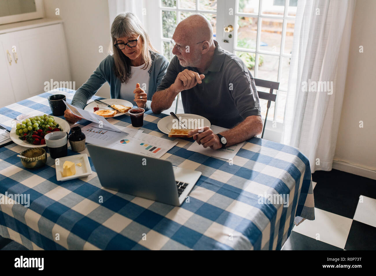 Senior man and woman having breakfast together at home with a laptop and papers on the table. Woman holding a paper discussing finances with her husba Stock Photo