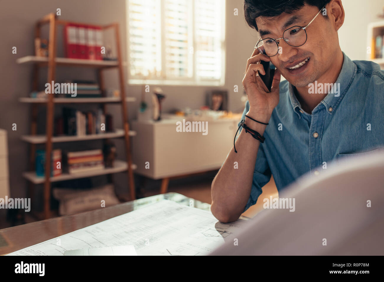 Happy architect looking at drawing and talking over cell phone at his home office. Architect answering phone call at his workplace. Stock Photo