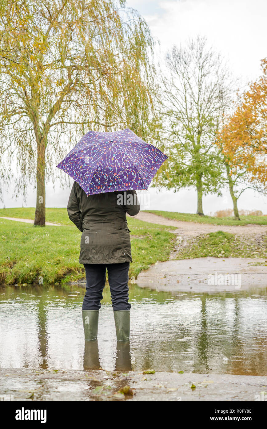 Kidderminster, UK. 7th November, 2018. UK weather: with today's persistent heavy showers (coupled with the significant overnight downpour), water levels are on the rise in Worcestershire with some areas experiencing flash flooding. A woman wearing wellies and holding an umbrella stands outdoors, rear view, isolated in rising water. Credit: Lee Hudson/Alamy Live News Stock Photo