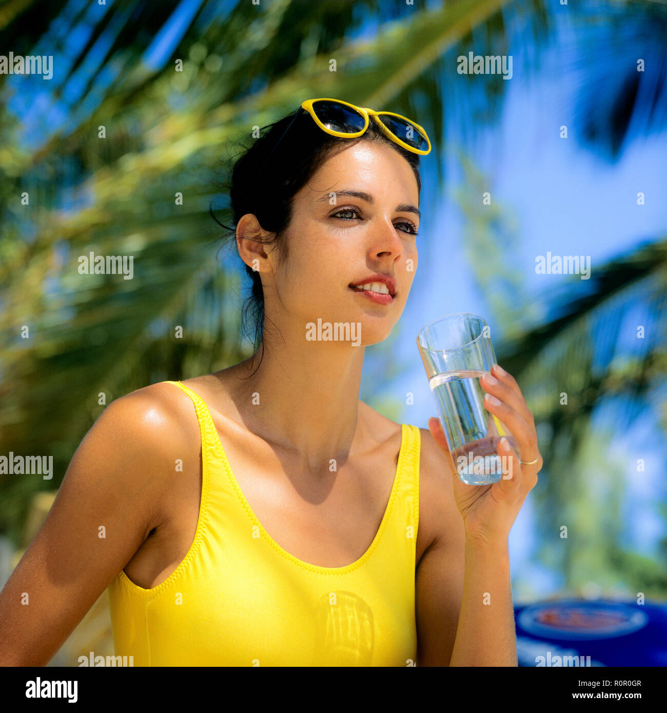 Young woman with yellow swimsuit holding a glass of water, palm trees background, Guadeloupe, French West Indies, Stock Photo