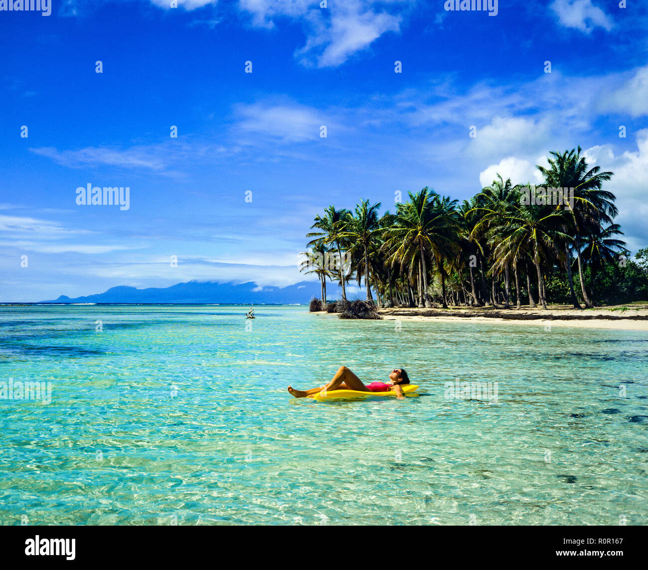 Young woman in red swimsuit sunbathing on yellow air mattress, floating on Caribbean sea, tropical beach, palm trees, Guadeloupe, French West Indies, Stock Photo