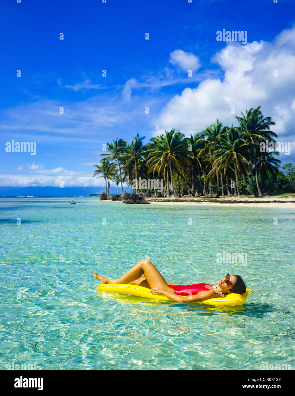 Young woman in red swimsuit sunbathing on yellow air mattress, floating on Caribbean sea, tropical beach, palm trees, Guadeloupe, French West Indies, Stock Photo