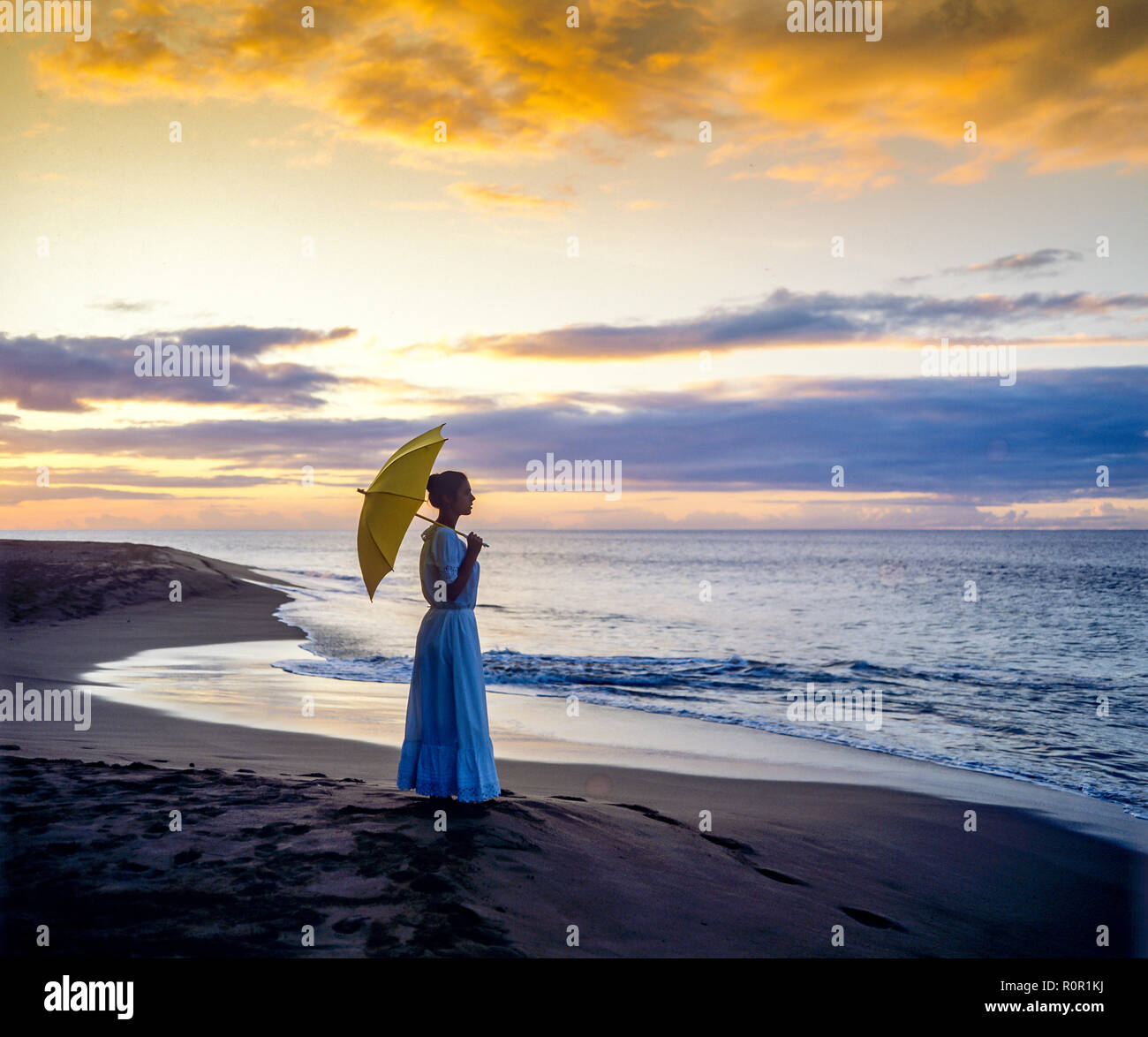 Young woman silhouette with long dress and umbrella facing the Caribbean sea at sunset, remote tropical beach, Guadeloupe, French West Indies, Stock Photo
