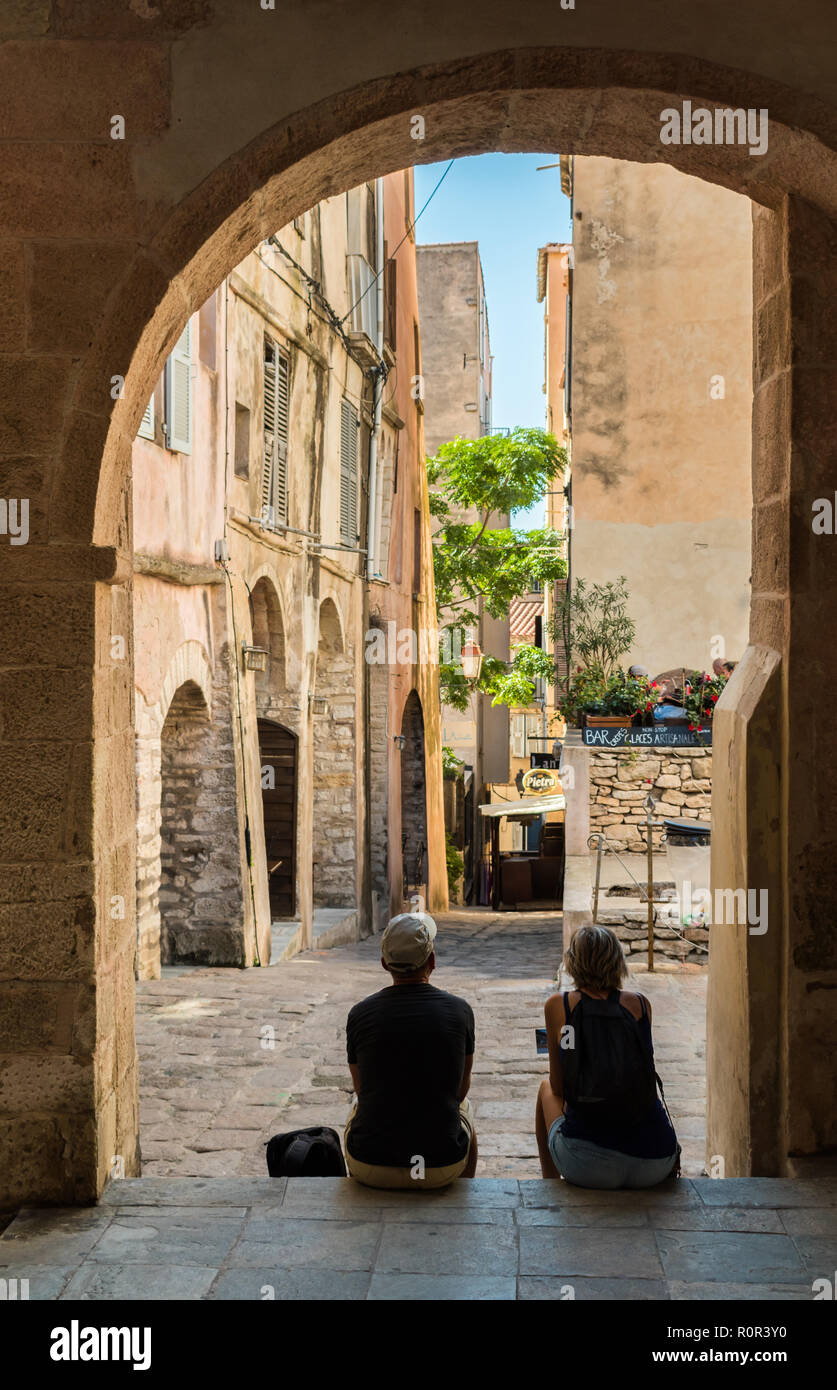 BONIFACIO, CORSICA – SEPTEMBER 30 2018: Two mature middle aged tourists take shelter from the hot midday sun by sitting on some steps. Stock Photo