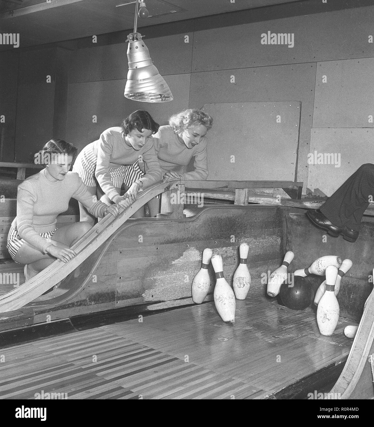 Bowling in the 1950s. Three girls are bowling for the first time ever. They are dressed in short striped skirts and jumpers and pictured at the area of the lane where the bowling pins are exactly in the moment the bowling ball hits a strike. The three girls are all theatre actresses; Ingrid Björk, UllaCarin Rydén and Brita Ulfberg. At this time this bowling alley have no machines that raises the bowling pins. Instead people were employed as pin raisers and the legs of such a person is seen to the right as he has his legs up in the air to avoid getting hit by the bowling ball or the pins.  1950 Stock Photo