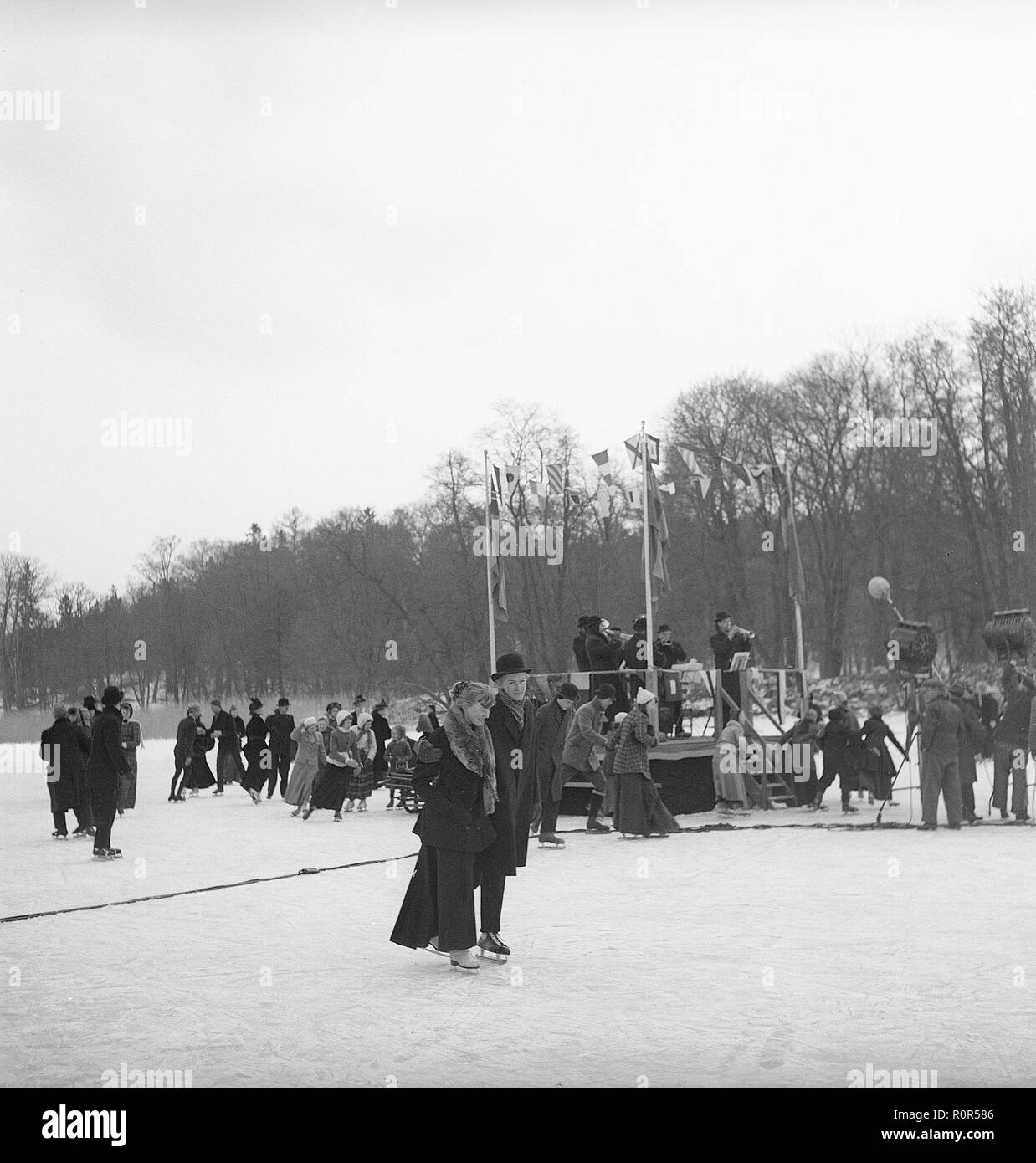 Figure skating at the turn of the century 1800-1900. A couple is pictured together when skating outdoors. In the background a typical swing sledge with children playing with it. Picture most probably taken on a film set. Sweden 1952. Photo Kristoffersson ref BF51-8 Stock Photo