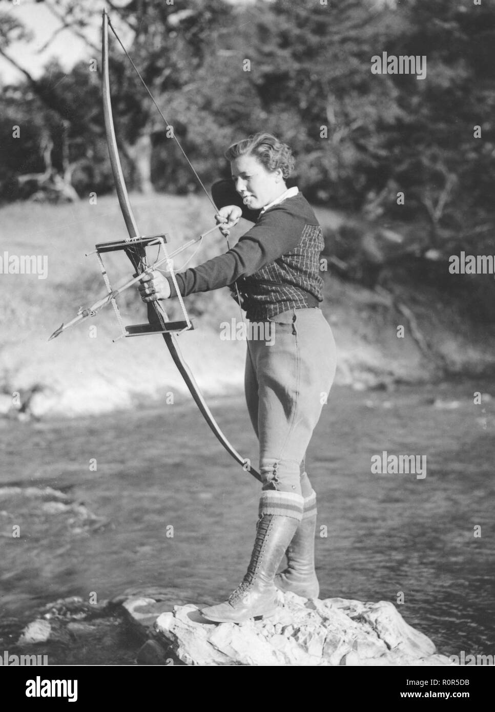 Women archer in the 1930s. A women uses her bow and arrow to catch fish and looks as if she is aiming to something in the stream. 1935 Stock Photo