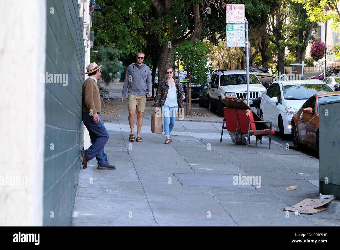 Male / female couple walk down the street as a man with a cigarette reclined against a wall observes them walking in his direction; street scene. Stock Photo