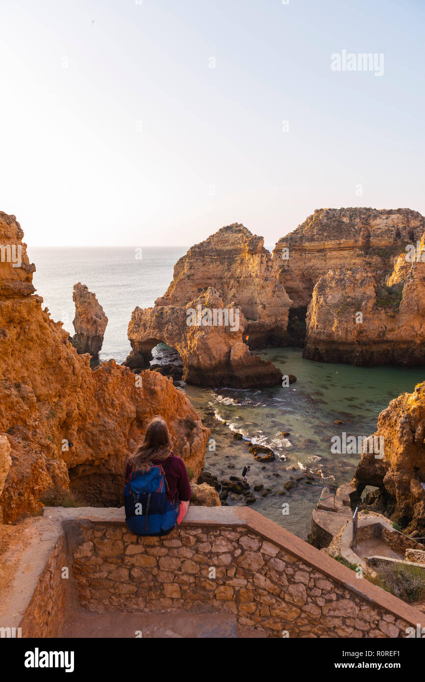 Female hiker sits on stone wall and looks over rocks in the sea, Algarve rocky coast, Ponta da Piedade, Lagos, Portugal Stock Photo