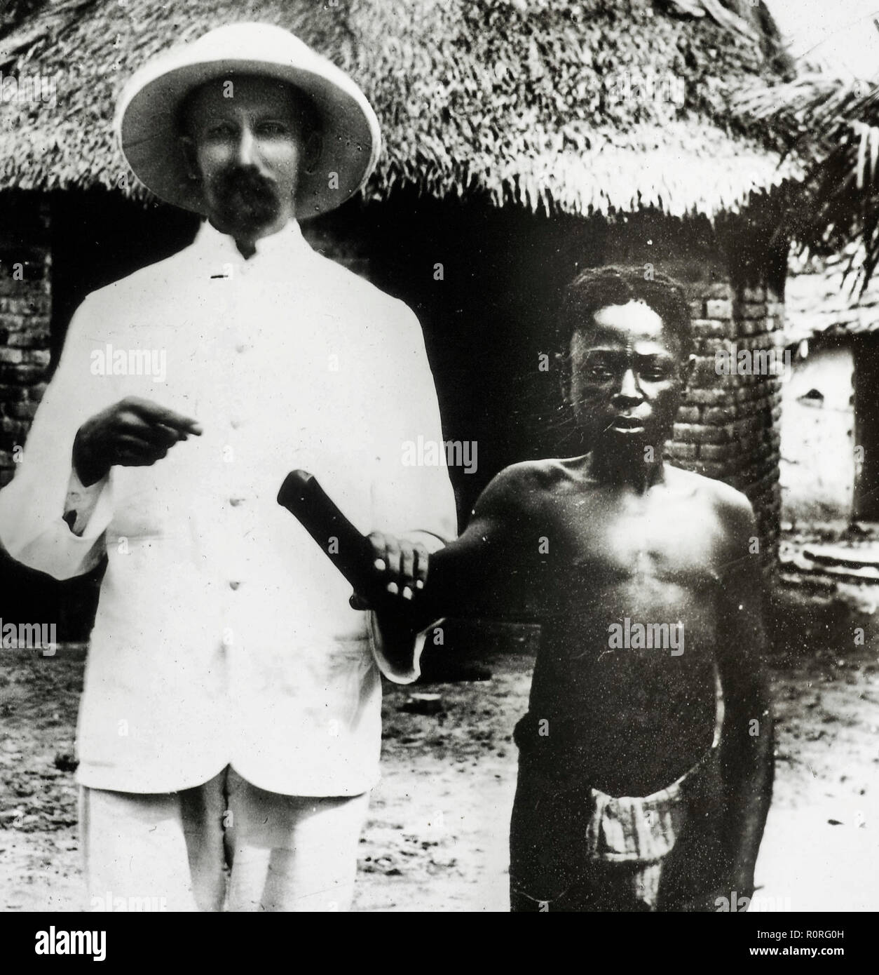 Black and white lantern slide showing a male missionary from the Congo Balolo Mission holding the arm of a Congolese man, Congo Free State (present day Democratic Republic of the Congo). The missionary is probably Mr Wallbaum, who wears a light safari suit and a pith helmet, and the Congolese man wears a loin cloth. The missionary holds up his companion's arm at the elbow, and points to the Congolese man's missing hand. The Congolese man is likely to have been a victim of the 'Congo atrocities', punishment, murders and mutilations (particularly amputation of the right hand on living victims or Stock Photo
