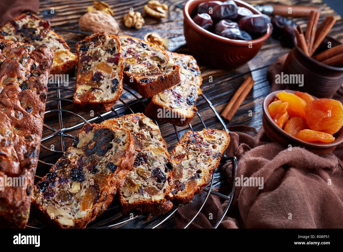 traditional dried fruits rich cake on a wire cake stand with brown cloth, cinnamon sticks, dried apricots and date fruits on a rustic wooden table, vi Stock Photo