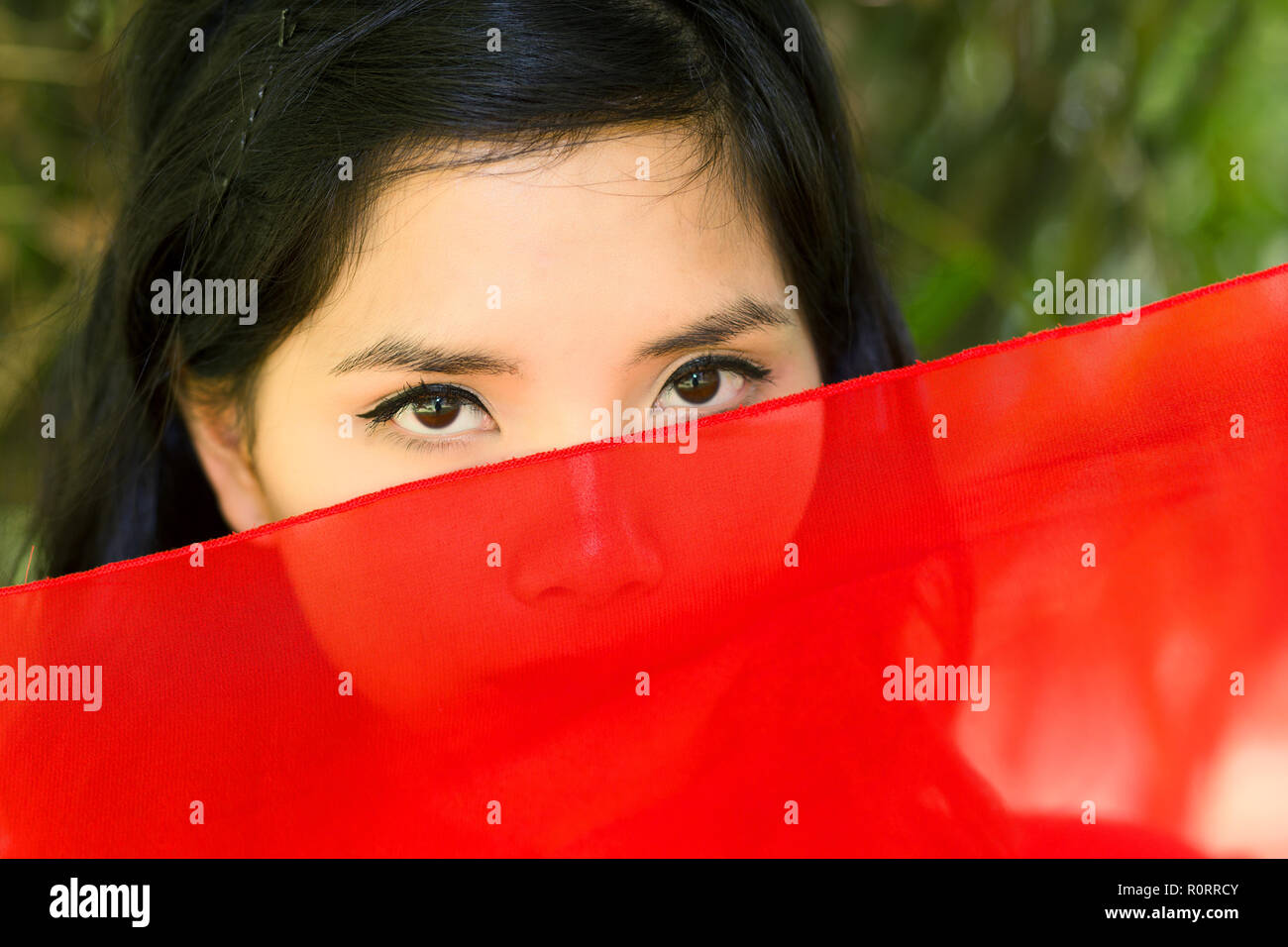 vietnamese woman peeking over red fabric at the camera so that just her eyes are visible, serious expression Stock Photo