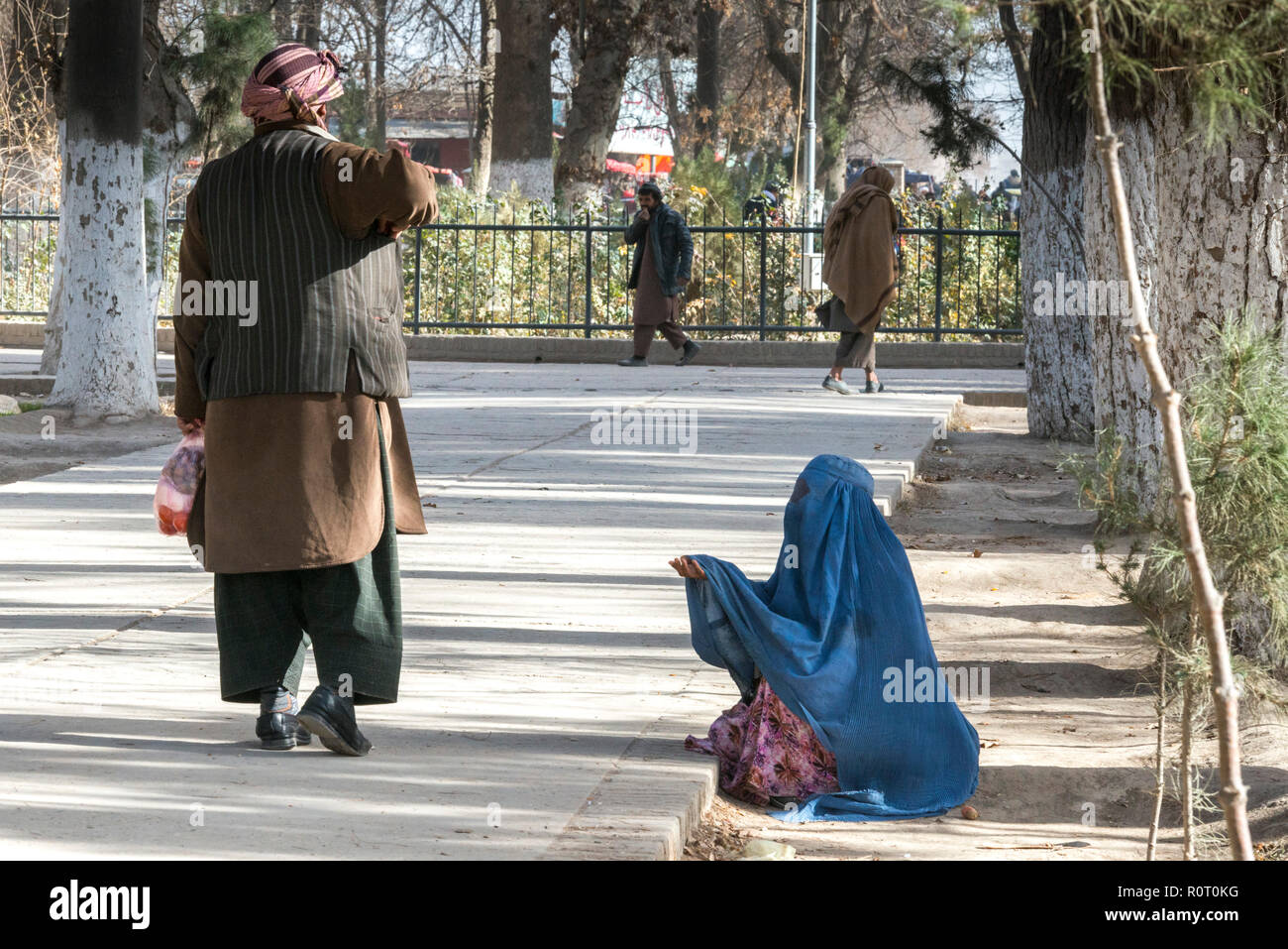 Lonely Hungry Female Beggar Wearing Traditional Blue Burqa Sitting On The Ground In A Park, Mazar-e Sharif, Balkh Province, Afghanistan Stock Photo